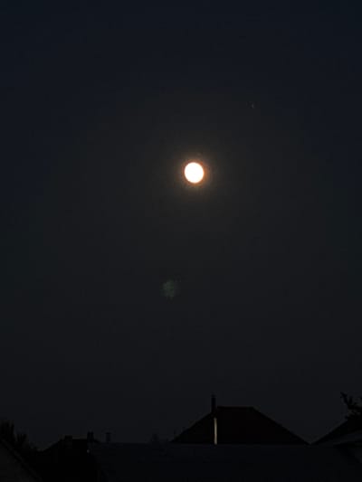 The image displays a night sky over Zniatsovo, Ukraine. A bright, luminous celestial body, likely the moon, is prominently featured in the upper center of the frame, casting a soft orange glow. Below it, a faint, green-tinted amorphous shape is visible, its nature unclear. The lower portion of the image shows the silhouettes of rooftops against the dark sky, with some structures appearing to have chimneys or attic vents. A single, small, bright point of light, possibly a distant star or satellite, is observable in the upper right quadrant of the sky. The overall impression is one of a clear night with celestial phenomena visible above the urban landscape.