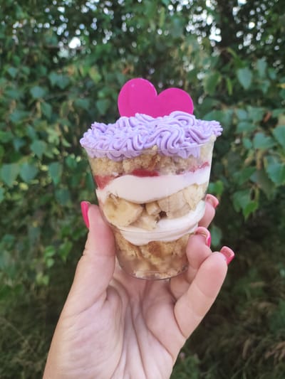 In a close-up shot, a hand with bright pink nail polish holds a transparent plastic cup containing a layered dessert. The dessert features alternating layers of light brown cake, white cream, sliced bananas, and a red fruit preserve. The top of the dessert is adorned with swirling purple frosting and a bright pink heart-shaped decoration. The background is a soft focus of green foliage, suggesting an outdoor setting, possibly a garden or park. The image was taken on the outskirts of the city of Myadzyel, Belarus.