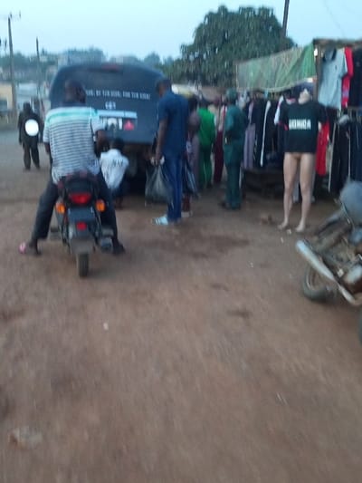 A bustling outdoor market scene unfolds. A motorcyclist waits near a black van with text on its rear. Clothes are displayed on a makeshift stand under a green tarp. Mannequins, one wearing a black shirt and the other without clothes, stand beside the rack. People mill around. Another motorcyclist approaches with their headlight shining. The ground is dirt and the sky is overcast.