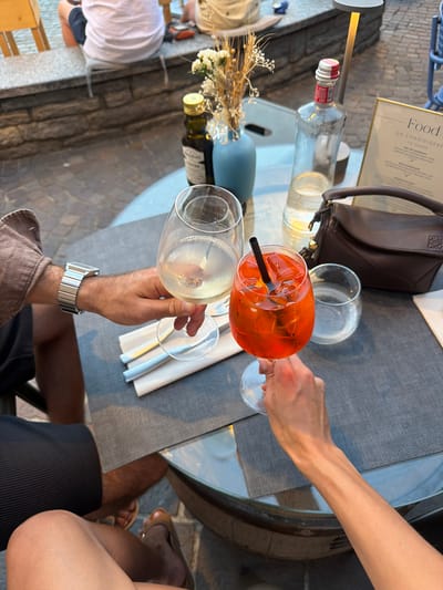 This image shows a close-up view of two people toasting at an outdoor table in Loveno, Italy. The table is set with drinks, including a glass of white wine and a bright orange aperitivo with a black straw. A brown leather handbag rests on the table next to a water bottle and a menu. In the background, people are seated at other tables, and a stone wall suggests a restaurant or cafe setting with a view of water. The overall impression is one of a relaxed, upscale dining experience in a scenic Italian location.