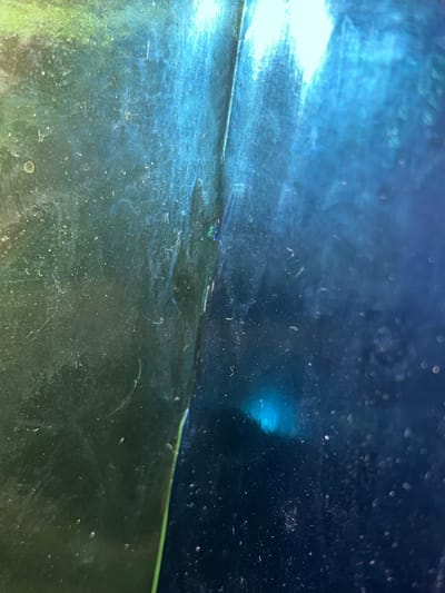 The image is an abstract, close-up shot dominated by shades of blue and green, focusing on a textured or reflective surface. A distinct, slightly irregular horizontal line traverses the image, creating a perceived division.

The upper portion of the frame is a deep, dark blue, filled with numerous small, scattered white specks and blurred circular light artifacts, resembling bokeh. A bright, light blue glow is visible in the upper right quadrant, suggesting a concentrated light source or reflection.

The lower portion of the image displays a mix of dark green and blue tones, with a similar scattering of white specks, streaks, and irregular patterns that could be dust, sediment, or water marks. A strong, bright white light illuminates a significant section of the lower left quadrant, creating a stark contrast with the darker areas. A thin, bright green line is faintly visible along the main horizontal division in some areas.

The overall impression is one of depth, light refraction, and surface texture, possibly an underwater view, a dirty pane of glass, or a macro shot of a complex material. There are no identifiable people, specific objects (beyond abstract textures), or discernible activities taking place. No text is visible. The image offers no specific cues to its stated location of Picassent, Spain.