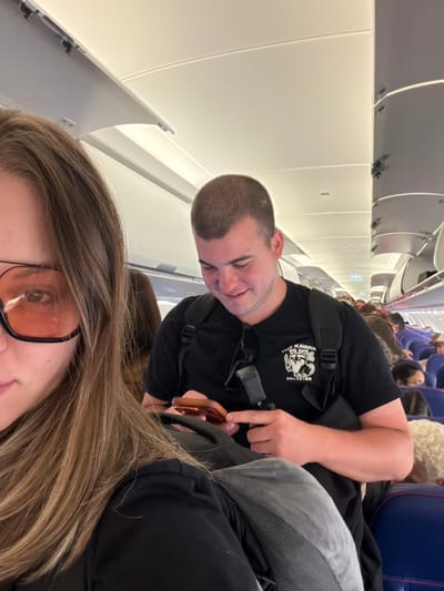 The image depicts a selfie taken inside an airplane cabin, likely at Orio al Serio, Italy, due to the visible livery on the seats and the overall interior design consistent with budget airlines operating from that airport. In the foreground, a woman with long, blonde hair and orange-tinted sunglasses is partially visible on the left. To her right, a man with short brown hair and a black t-shirt with a graphic print is looking down at his phone. He is wearing a backpack. Other passengers are visible in the rows behind them. The lighting suggests the cabin is illuminated by the overhead lights. The scene captures a moment during a flight, with people seated and engaged in personal activities.
