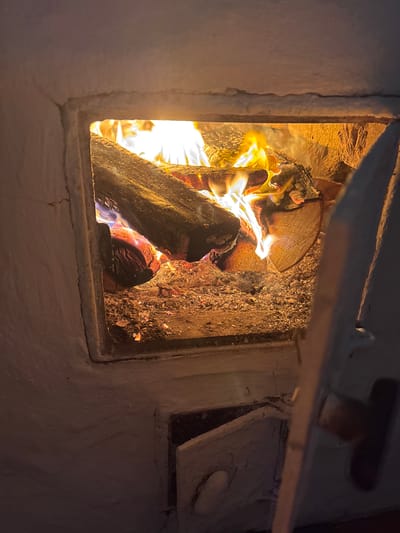 The image shows a close-up view of a burning fireplace or stove, likely made of white plaster or brick. Logs are actively burning inside, with visible flames, embers, and ash. The opening of the fireplace is rectangular. To the lower right, a small, open metal door, possibly for ash removal, is visible. The scene is dimly lit, with the primary light source being the fire itself. The image does not contain any information about the city of Курган, Russia, or any people.