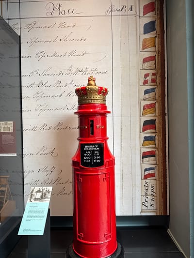 The image shows a red pillar box with a crown on top, positioned in front of a large display featuring handwritten text and small illustrations of flags. The pillar box has a plaque that reads "HOURS OF COLLECTION" with a schedule below it. To the left of the pillar box, there is an information panel with text and an illustration of an early pillar box. In the background, a large, weathered document with calligraphic handwriting and a series of small, colorful flags arranged vertically is visible. The overall setting appears to be a museum exhibit. The specific location in the City of Edinburgh, United Kingdom is the National Museum of Scotland.