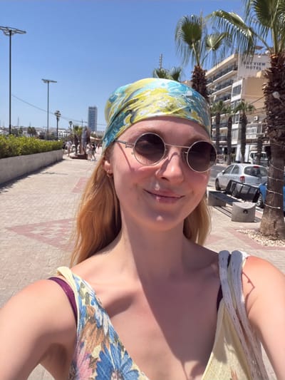 This is a slightly overexposed, vertical, selfie-style photograph taken outdoors on a sunny day. The main subject is a young woman with blonde hair, wearing round, reflective sunglasses and a blue and yellow patterned headscarf. She is also wearing a yellow top with a floral pattern and a dark purple bikini bottom. Her expression is a smiling, relaxed one, suggesting a happy and casual mood.
The setting appears to be a seaside promenade or walkway. In the background, there are palm trees, a building with "BAY VIEW HOTEL" visible on its facade, a white car, and streetlights. The ground is paved with light-colored rectangular tiles, some of which have red diamond-shaped accents. The sky is a clear, bright blue, indicative of good weather and likely daytime, possibly late morning or afternoon given the strong sunlight and shadows.
The overall impression is one of a tourist enjoying a warm, sunny day in a coastal resort town. The image captures a moment of leisure and enjoyment.