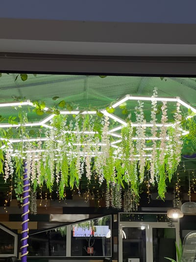 In the city of Shchyolkine, Ukraine, an interior scene is captured, likely within a cafe or restaurant, as suggested by the reflective surfaces of windows showing a glimpse of the outdoors and interior lighting. The focal point of the image is a hexagonal light fixture suspended from the ceiling, adorned with a profusion of artificial white and pink floral cascades and green foliage. These hanging decorations create a lush, cascading effect, reminiscent of wisteria.
Integrated into the hexagonal structure are bright, linear LED lights, illuminating the artificial flora and casting a soft glow. Beneath this main display, a series of delicate, warm-toned fairy lights are strung, adding a whimsical ambiance.
To the left, a prominent purple pillar wrapped in beige rope serves as a vertical element within the composition, further enhancing the decorative aesthetic. The background consists of a dark wall with horizontal paneling in various shades of wood and black, punctuated by rectangular reflective surfaces.
The overall impression is one of a thoughtfully decorated, possibly whimsical, indoor space, designed to create an inviting and visually engaging atmosphere. The image appears to be taken at dusk or night, given the exterior darkness visible through the windows.