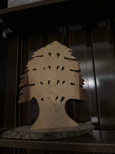 A wooden fish-shaped object is displayed on a dark, horizontal slatted surface, likely a shelf or wall. The fish is crafted from light-colored wood and has numerous small, oval-shaped holes cut into its body. The edges of the fish are rough and irregular, giving it a rustic appearance. A larger piece of wood with a natural, bark-covered edge is attached to the right side of the fish, extending outward like a tail or fin. The background consists of dark, horizontally oriented wooden slats that appear to be polished and slightly reflective, suggesting an indoor setting. The lighting is dim, casting shadows and highlighting the texture of the wood. The overall scene evokes a sense of handcrafted decor, possibly for a home or a workshop. No people, text, or specific weather conditions are visible. The location context of Pakuro, Nigeria, does not provide any directly observable cues within the image itself.