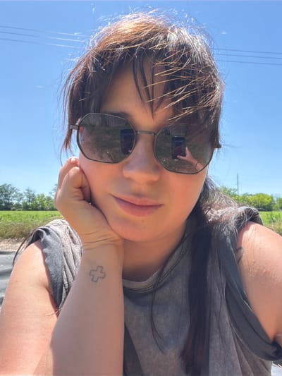 A young woman is taking a selfie outdoors on a sunny day. She is wearing sunglasses and resting her chin on her hand. Her hair is dark and slightly messy. She is wearing a gray t-shirt. There is a small cross tattoo on her left wrist. The background is a blue sky with some power lines and trees. The location is not specified.