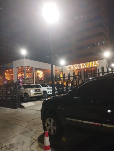 The image depicts the exterior of "Casa Tarbes," a restaurant in Valencia, Venezuela, at night. The restaurant's facade is illuminated with warm lighting, highlighting its name in large letters. In front of the restaurant, several cars are parked, including a black Grand Cherokee prominently featured in the foreground and other tan and white vehicles partially visible. A red and white traffic cone sits near the front wheel of the black SUV. The building behind the restaurant appears to be a multi-story structure with lights on some floors. Streetlights cast a bright glow over the scene. A wrought-iron fence partially obscures the view of the restaurant. The sidewalk is visible in the foreground.