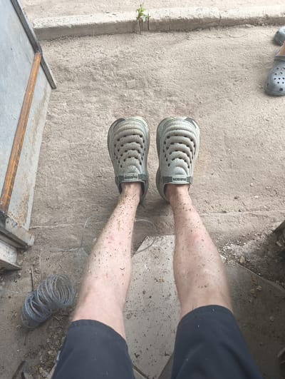 In the city of Rassvet, Kazakhstan, a person's legs are shown from a high-angle, downward perspective. They are wearing light gray, perforated clogs with a black strap that says "FASHION". The legs are covered in small bits of green debris, possibly grass or leaves. One leg has a few red marks, suggesting minor abrasions. The background is a concrete or asphalt surface, with a rusted metal structure on the left and a coiled wire on the ground to the left of the legs. A small green plant is visible growing from a crack in the concrete in the upper background.