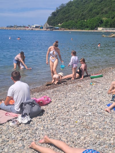 This is a sunny outdoor photograph taken at the beach in Dzhubga, Russia. Several people are present, some in the water and others on the pebbled shore.
In the foreground, a man wearing a white t-shirt and blue denim shorts is sitting on the pebbles, facing away from the camera. Next to him is a gray backpack, a pink inflatable flamingo, and a pair of white sneakers. Further down the shore, a person's legs are visible, wearing blue and white patterned swimwear.
In the middle ground, on the edge of the water, a woman in a white bikini is standing and looking down. To her left, a woman in a pink shirt and denim shorts is in the shallow water, interacting with a child in a white swimsuit. Another child, wearing a sun hat, is crawling on the pebbles near the water's edge. A woman in a black swimsuit is sitting on the pebbles to the right of the children. A green inflatable watermelon slice toy is next to her.
In the background, the blue sea stretches out, with a few people swimming or wading. A rocky breakwater with several buildings on it juts into the sea. Beyond the water, a tree-covered hillside rises steeply. The sky is bright blue with a few scattered clouds. The overall impression is of a leisurely summer day at the beach.