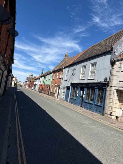 This media file presents a sunlit street scene in Gorleston-on-Sea, United Kingdom, captured from a slightly low perspective looking down a row of buildings. The clear, bright blue sky is adorned with scattered, wispy white clouds, indicating fair weather. Long, sharp shadows stretch across the asphalt road, suggesting a time of day with a low sun, either morning or late afternoon.
On the left side of the frame, a succession of terraced buildings lines the street. In the foreground, a red brick building with two black satellite dishes on its roof is visible. Adjacent to it stands a prominent light blue-grey building featuring white-framed windows and a dark blue shopfront. This establishment displays clear text on its fascia: "GORD BLUES" and "Open Late" on the left, and "Spirits," "Real Ales," "Good Food" on the right. Further down the street, a variety of colorful buildings, including green and red brick facades, are visible, with a distinctive building featuring a clock face in the mid-distance.
The street itself is an asphalt road with double yellow lines running parallel to the curb on the left. A significant portion of the road on the right is covered by a deep, dark shadow, contrasting with the sunlit section and highlighting the intensity of the sunlight. No people or specific activities are visible, lending a quiet atmosphere to the street. The architectural style of the buildings is characteristic of a traditional British town or coastal area.