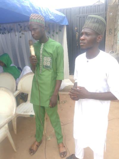 Two adult males stand outdoors on reddish-brown earth in Ijoko, Nigeria. The male on the left wears a green traditional tunic and trousers with white and green embroidery, a brown and beige patterned skullcap, and brown sandals. He holds a gold-colored microphone in his right hand. The male on the right wears a white traditional tunic and trousers with a dark green patterned skullcap; his hands are clasped in front of him. In the left background, three white plastic chairs are visible, with a person in a green hijab seated behind them. Above them, a blue tarpaulin canopy is draped with white fabric. The right background features a dark corrugated metal gate and a light-colored wall with faint markings.