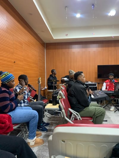 A band plays music indoors. A man sings into a microphone while seated on a red chair. A drummer plays a drum set. A man plays a bass guitar. The band is playing for a group of people sitting on chairs. The space has a wood-paneled wall. A television hangs on the wall behind the band.