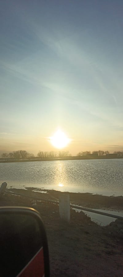 This image displays an outdoor landscape scene at sunrise or sunset, located in Xojeli, Uzbekistan. The upper portion of the frame features a clear sky, gradually transitioning from a light blue hue at the zenith to a warm yellow-orange closer to the horizon. The sun, a brilliant, intense light source, is positioned centrally just above the horizon, casting a strong glare and reflecting vibrantly across the water.
The mid-ground is dominated by a wide body of water, possibly a lake or large canal, its surface exhibiting ripples that reflect the sun's light in a shimmering pattern. A distant treeline, composed of bare deciduous trees, extends horizontally along the far bank, silhouetted against the bright sky.
In the immediate foreground, a dark, uneven dirt or muddy bank is visible along the water's edge on the right. Two light-colored, weathered vertical posts protrude from this bank. A dark, horizontal pipe or bar runs parallel to the bank. The lower-left corner of the image is partially obscured by a dark, curved object, identifiable as a vehicle's side mirror, which displays a distinct red stripe along its lower edge. There are no individuals or discernible actions present in the scene.