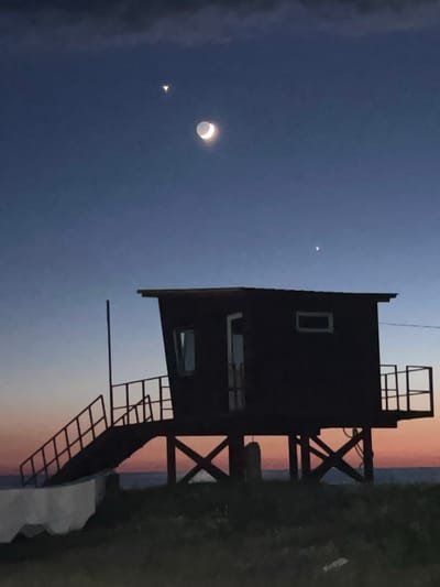 An image taken in Shekvetili, Georgia, depicts a silhouette of a small wooden structure, possibly a lifeguard station or a small lookout, against a twilight sky. The structure is elevated on stilts and features a staircase leading up to its entrance. The sky transitions from a deep blue at the top to a warm orange and pink hue near the horizon, indicating either sunrise or sunset. In the sky, a crescent moon is visible, along with two bright celestial objects, likely planets, positioned above and to the right of the moon. The foreground is a grassy area with some indistinct objects. The overall scene is serene and atmospheric.