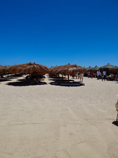 This is a medium-shot, eye-level photograph of a sandy beach with thatched umbrellas and several people. The sky is a clear, vibrant blue. The sand is light brown and appears to be a fine grain, with some footprints visible. In the foreground, the sand takes up the majority of the frame. Scattered across the middle ground are several thatched umbrellas providing shade. Under one umbrella, two women are sitting and facing each other, possibly conversing. Behind them, more umbrellas are visible, and a few people are standing or walking around. In the background, more umbrellas and people are present, creating a lively beach scene. The overall impression is a sunny day at a resort in Hurghada, Egypt.