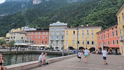This media file captures a vibrant scene in Riva del Garda, Italy, with the image rotated 90 degrees counter-clockwise. In the foreground, several people are walking along a cobblestone promenade. One individual, wearing a white t-shirt and blue shorts, walks away from the viewer. Another person, in a white t-shirt and green shorts, walks in the same direction. A third person, also in a white t-shirt, stands with their back to the camera.

Across a reflective body of water, a row of colorful buildings lines the waterfront. The buildings exhibit a European architectural style, with multiple stories, arched windows, and balconies. The colors range from a bright orange to yellow and blue. Visible text on one of the buildings reads "HOTEL EUROPA".

In the background, lush green hills rise steeply, indicating a mountainous or hilly terrain surrounding the town. The water appears calm, reflecting the buildings and the sky. A few umbrellas and outdoor seating areas suggest cafes or restaurants. The presence of trees on the hills and a cable car system traversing the slopes hint at a scenic and possibly tourist-oriented location. The overall atmosphere appears to be daytime, with good weather, likely a sunny day given the brightness of the scene. The mood is relaxed and leisurely, with people enjoying the waterfront promenade and the picturesque views.