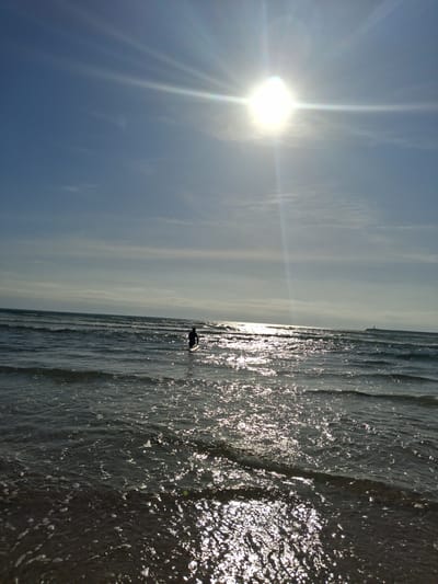 The image shows a beach scene in Figueira da Foz, Portugal. The bright sun is in the upper left of the frame, with light rays extending across the sky. The sky is blue with some clouds. The Atlantic Ocean occupies the right side of the image, with waves reflecting the sunlight. Near the center of the image, a person in dark attire is visible in the water. The beach is dark sand.