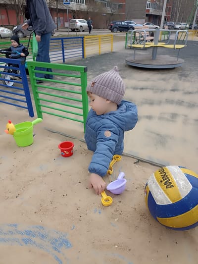 A child in a blue coat and gray hat is playing in a sandbox. The sandbox contains several plastic toys, including a green bucket, a red bucket, and yellow and purple shovels. A Mikasa volleyball is also in the sandbox. Another child is sitting in a stroller pushed by an adult. A playground with a merry-go-round is visible in the background, along with cars and buildings. The location is Serpukhov, Russia.