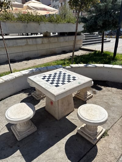 In the city of Donji Pobori, Montenegro, a public outdoor space features a stone chess table with a built-in chessboard. Two matching stone stools are positioned around the table, which is set on a concrete ground. A low, curved concrete wall surrounds the area, with grass and small trees growing behind it. In the background, there are more trees, a building, and a set of concrete stairs. The scene is bathed in sunlight, casting distinct shadows. The chess table has some graffiti markings on its surface.