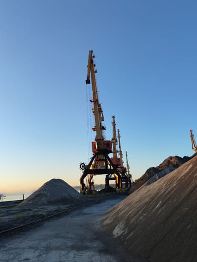 This is a low-angle, full shot of a port facility under a clear blue sky at sunrise. The image features several large, yellow and red construction cranes, their booms extended upwards. In the foreground, there are large piles of gravel and sand. Train tracks run between the piles and the cranes. In the background, there is a body of water and a hazy shoreline. The location is the port of Bol'shie Mkhi, Russia. There are no people or animals visible in the image. The overall scene is industrial and barren, with the rising sun casting long shadows and highlighting the textures of the materials.