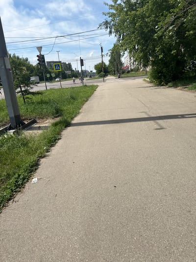 The image depicts a street scene in Angarsk, Russia, on a bright, partly cloudy day. A paved sidewalk runs diagonally across the foreground, bordered by a grassy verge on the left. In the distance, a traffic light with a red signal is visible, along with pedestrian crossing signs. Several overhead wires crisscross the sky. A line of trees and buildings can be seen along the street. Two individuals are walking in the distance, and a white truck is parked to the left. The overall impression is of a typical urban environment.