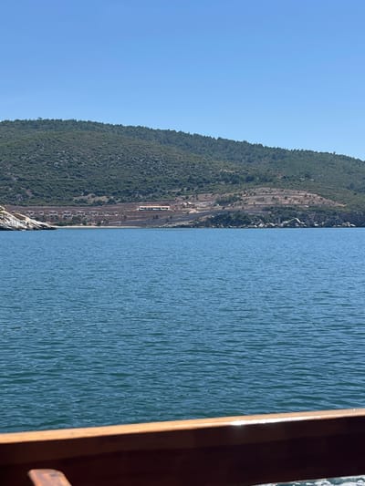 The image displays a clear coastal landscape under a bright, cloudless blue sky. The scene appears to be taken from the water, possibly from a boat or pier, as indicated by a wooden railing or structure visible along the far right edge of the frame.

The right portion of the image is filled with the tranquil blue-green waters of what is likely the Aegean Sea, exhibiting gentle ripples that catch the sunlight. To the left, a prominent, heavily forested hillside or mountain range stretches horizontally, its slopes densely covered with green trees and some visible rocky outcrops at its base.

Running parallel to the coastline and nestled at the foot of the hillside is a railway line. Multiple tracks are visible, and several light-colored train carriages or an entire train can be seen on one of the tracks, appearing to be stationary or moving slowly. Some smaller buildings or structures are also discernible adjacent to the railway. The railway line forms a distinct man-made ribbon between the natural elements of the water and the forested land.

The upper left section of the image shows a vast expanse of unblemished blue sky, signifying clear and sunny weather. The bright illumination across the scene suggests it is daytime, likely mid-morning or early afternoon. No individuals are visible in the frame, and no specific human activity is overtly depicted, though the presence of the train implies transport operations. The setting is a scenic coastal area near Menderes, Turkey, characterized by the dramatic interaction of mountains, sea, and human infrastructure. There is no visible text in the image.