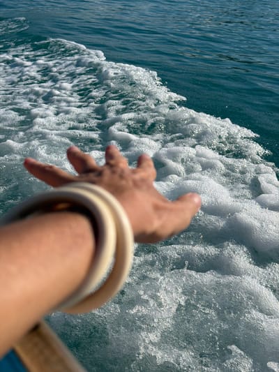 This media file captures a dynamic perspective of a hand reaching into frothy, turquoise water, likely from a moving vessel. The scene is dominated by the churning wake of a boat, creating a texture of white foam and sparkling water droplets. The hand, adorned with two light-colored bangles on the wrist, is blurred due to motion, suggesting the speed of the water or the camera. The fingers are outstretched, seemingly about to touch or interact with the water.
The setting appears to be a bright, sunny day, with the sunlight glinting off the water and foam, creating a sense of energy and movement. The clear, deep turquoise of the undisturbed water to the left contrasts with the white, effervescent wake, highlighting the motion. This perspective offers a sense of immersion and the exhilaration of being on the water. The location context of Baan Thong Krut, Thailand, evokes a tropical marine environment. No text is visible in the media file. The overall mood is one of active engagement with the surrounding natural elements.