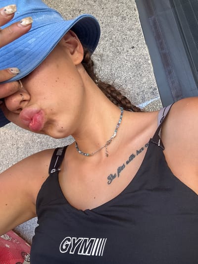 This is a close-up, vertically oriented photograph of a young woman lying on a textured, grey surface, possibly concrete or asphalt. She is wearing a black sports bra or athletic top with white lettering that reads "GYM" on the right side and appears to have a grey and black striped band along the top edge. A thin, beaded necklace with blue and silver beads is visible around her neck.
The woman's face is partially obscured by a light blue baseball cap worn backward. Her lips are puckered, and her right hand, with painted nails, is near her face. A small silver ring is visible on her right index finger. She has a tattoo on her collarbone that appears to read "She flies with her O". Her hair is styled in a braid that falls over her shoulder.
The lighting suggests it is daytime, with natural light illuminating the scene. The background consists of the grey surface and a dark grey, textured object, possibly a railing or grating, running horizontally across the top of the image. The overall impression is a casual, outdoor setting. There are no discernible emotional cues beyond the facial expression of puckered lips.