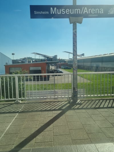 The image shows an outdoor scene at the Sinsheim Museum/Arena in Sinsheim, Germany. The scene is composed of a sign indicating the location, a metal fence, a gray stone wall, a green grassy area, and buildings in the background. The sky is bright blue. The sign reads "Sinsheim Museum/Arena" vertically in white letters on a dark blue background. The metal fence casts a shadow on the gray stone wall. In the background, there are buildings with unique architecture, including a large hangar-like structure, as well as cars and other vehicles. There are no people visible in the image.