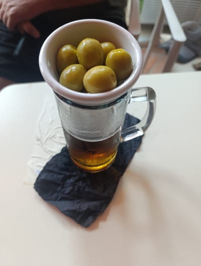 A close-up, eye-level shot shows a clear glass mug filled with beer, condensation on its exterior, sitting on a white table. The mug is partially obscured by a crumpled black napkin. A small white dish, containing several green olives, is perched on top of the mug, appearing to be part of a serving presentation. In the background, a person's left arm and shoulder are visible, with dark fabric suggesting clothing. The setting appears to be an outdoor or indoor seating area, possibly a bar or restaurant, given the presence of the drink and the table. The lighting is natural, and the overall composition focuses on the beverage and olives. The image was likely taken in Águilas, Spain, a coastal town known for its bars and restaurants.