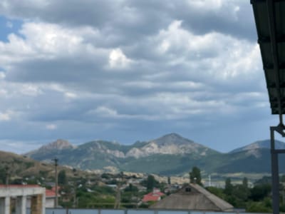 The image captures a cloudy sky over a village situated in front of a mountainous landscape. In the foreground, on the right, a dark metal structure, possibly a part of a roof or a canopy, is visible. On the left, a portion of a building with exposed concrete pillars and a white façade is present. The middle ground reveals a residential area with houses, some featuring red roofs, nestled amongst green trees and hills. Utility poles are scattered throughout the village. The background is dominated by rugged mountains, with a prominent peak in the center. The overall scene depicts a rural or semi-rural environment under an overcast sky. The location is identified as within the Shchebetovsky settlement council, Ukraine.