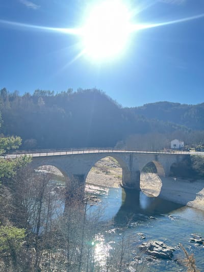 The image displays a multi-arch stone bridge spanning a river under a clear, intensely bright blue sky, with the sun prominently centered in the upper frame, generating significant lens flare. This scene is located in Stoianovo, Bulgaria. The bridge, constructed of light-colored masonry, features a wooden handrail along its top and extends across a river with visible shallow areas, rocky banks, and sandbars. The river water is clear, reflecting sunlight and sky. On the left side, evergreen foliage and bare deciduous trees are visible in the foreground, with densely forested hills rising in the background. On the right side of the bridge, a small, white, single-story building stands on the riverbank, accompanied by additional green structures further along the bank. No individuals, vehicles, or animals are present.