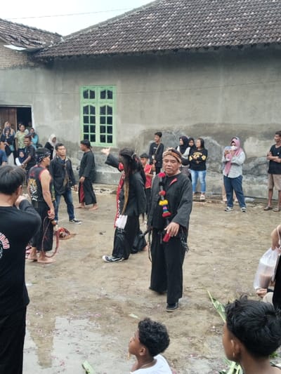 The scene depicts an outdoor cultural or ceremonial event taking place in the city of Kerjo, Indonesia. The immediate background features a two-story building constructed with unpainted light gray concrete walls and a dark brown, terracotta-tiled roof. A bright green window with multiple panes is centrally located on the upper portion of the wall, and an open doorway on the left reveals several observers inside.
The ground is an uneven, damp, light brown earth surface, showing puddles of water and scattered pink flower petals and green leaves. The sky above is overcast and bright.
In the mid-ground, a group of individuals appears to be participating in or performing an activity. Prominently featured are two men in black attire. One man, facing right, wears a black long-sleeved shirt, black pants, and a patterned head covering, with a colorful beaded necklace (yellow, red, green, blue elements) hanging from his neck. He holds a red object, possibly a handle or part of a whip, in his left hand. To his left, another participant wears all black clothing, including loose-fitting pants and a long-sleeved top, and has long dark hair, red facial markings, and a white sash or small bag across their torso. This individual is wearing black converse-style sneakers and has their right arm raised and hand extended in a gesture.
To the left of these two, another man in a black sleeveless vest with a tiger graphic on the back is visible from behind, holding a braided red/brown whip-like object. He is barefoot, as is another man next to him wearing a black t-shirt and blue jeans. In the immediate foreground, two young boys are present; one in a white t-shirt looks upwards and to the right, while the other with dark, curly hair is partially visible.
A larger group of onlookers is situated in the background and right side of the frame. This group includes several women, some wearing hijabs, and young men. One woman in a pink long-sleeved shirt and blue jeans is holding a white smartphone horizontally, appearing to record the event. Another individual on the far right holds a transparent plastic bag containing a dark liquid.