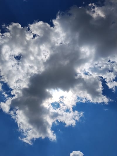 A low-angle, portrait-oriented shot captures a dynamic sky with a bright blue expanse dominating the upper portion. Scattered across the sky are numerous clouds, varying in size, density, and color. Towards the center, a large, dark cloud casts a shadow, creating a dramatic contrast with the sun that peeks through a gap in its lower section, emitting a bright, diffused light. Lighter, wispy clouds surround this central mass, some catching the sunlight and appearing brilliant white, while others are tinged with gray. The overall impression is one of a dramatic and ever-changing sky, typical of a day with mixed weather conditions. The image contains no discernible elements that would indicate a specific location within the city of Hoholeve, Ukraine.