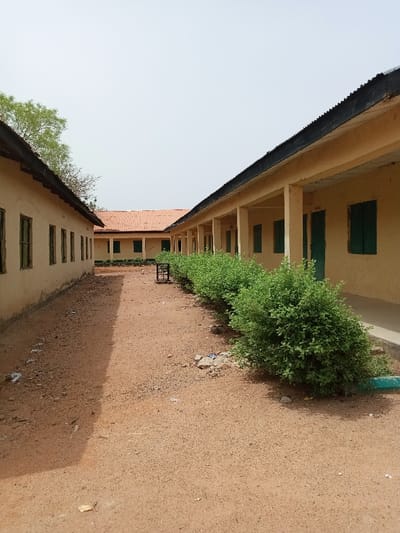 The image shows an outdoor courtyard between two buildings. The buildings are both painted a pale yellow and have open verandahs. The verandah of the building on the right side of the image has green window shutters and doors. There are green bushes planted along the edge of the veranda. The buildings are made of concrete blocks. The courtyard is covered in a brown, sandy soil, with a few scraps of litter. There are no people in the image. The location, based on the coordinates provided, is Rumde Waziri, Nigeria. The image appears to have been taken during the day.