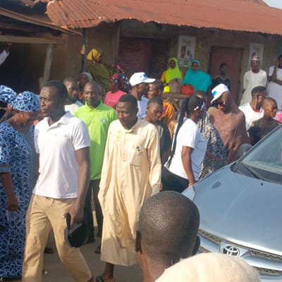 A crowd of people congregates in Gudi, Nigeria. Individuals wearing casual attire and traditional clothing are present. A light-colored car is partially visible on the right. In the background, a building with a corrugated metal roof is noticeable. Political posters are adhered to the wall of the building. The environment is outdoors, with natural light illuminating the scene.