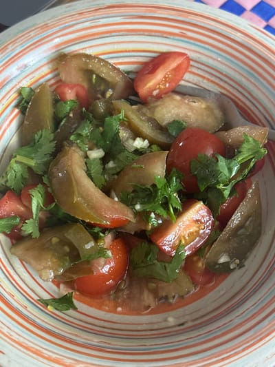The image displays a vibrant, close-up overhead view of a fresh tomato salad or salsa served in a ceramic bowl. The bowl features a distinctive pattern of red, orange, blue, and white stripes swirling outwards from the center.

The dish primarily consists of various types of chopped and sliced tomatoes. Bright red, halved cherry or grape tomatoes are interspersed with larger slices of darker, possibly heirloom or beefsteak tomatoes that exhibit a brownish-green hue. Generous amounts of fresh, green cilantro leaves are scattered throughout the salad, along with small pieces of what appears to be minced garlic or onion, adding texture and flavor. The tomatoes appear fresh and juicy, with some liquid visible at the bottom of the bowl. A silver utensil, possibly a spoon, is partially submerged in the upper left portion of the dish.

The scene captures a prepared dish, suggesting an upcoming meal or a recently served side. There are no people visible, and no specific time of day or weather conditions can be determined from the image. No text is visible in the frame. The dish presents as a fresh, homemade accompaniment, fitting for a meal in Egkomi Lefkosias, Cyprus.