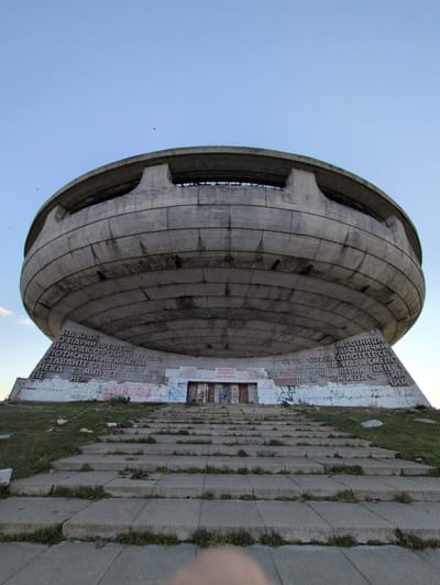 This is a vertical, eye-level, wide shot of the Buzludzha monument, a derelict communist-era monument in Bulgaria. The concrete structure, a large disc-shaped building, dominates the left side of the frame, with its ribbed surface and circular opening visible. To the right, a long, stepped concrete pathway ascends, overgrown with grass in places. The pathway is flanked by a white concrete wall covered in graffiti and large Cyrillic text, which appears to be slogans or historical inscriptions.
The sky is a clear, pale blue, suggesting a bright, possibly daytime setting, though the angle of the sun is not readily apparent. The monument itself shows signs of age and neglect, with some sections appearing weathered and covered in what might be dirt or lichen. The graffiti adds a layer of modern decay to the historical structure.
The visible text on the wall includes phrases in Cyrillic script, such as "РАБОТНИЦИ РАБОТНИЧКИ" (Workers, female workers) and "СПЛОТЕТЕ СЕ НАПРЕД ДРУГАРИ" (Unite forward comrades). There are also signs indicating "ВЛИЗАНЕТО ЗАБРАНЕНО!" (ENTRY PROHIBITED!), suggesting the monument is closed to the public. The overall atmosphere is one of grand historical ambition now fallen into disrepair, evoking a sense of both past power and present abandonment.