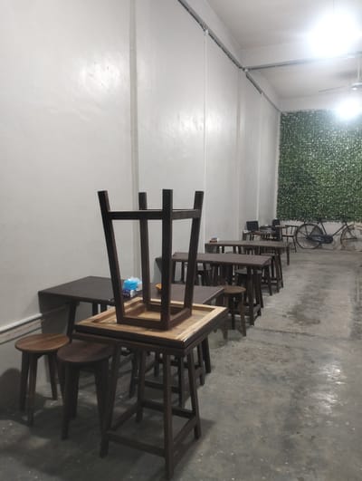 The image depicts the interior of a cafe or eatery in Tingkeummanyang, Indonesia. The scene is set in a long, rectangular room with a concrete floor and white, textured walls. The lighting suggests it is either evening or an indoor space with artificial illumination. Several wooden tables and chairs are arranged in rows, most of which are empty and appear to be set up for customers. In the foreground, a table has a chair placed upside down on it, and a packet of tissues is visible. Further back, a row of tables is accompanied by small, round stools. Towards the far end of the room, one table has a laptop and other items, suggesting a workspace or a different section of the establishment. A prominent feature in the background is a wall covered in artificial green foliage, creating a naturalistic contrast with the otherwise minimalist interior. To the right of the foliage wall, a vintage-style bicycle is parked. There is no visible text in the image. The overall atmosphere is quiet and empty, implying the establishment is either closed or between busy periods.