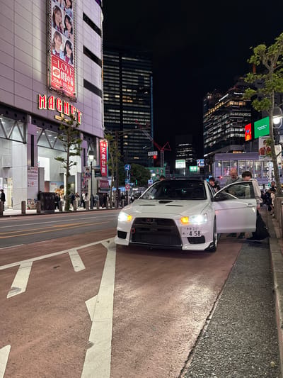 The image depicts a nighttime scene in Shibuya, Japan, centered around a white car parked on the side of a road. People are seen around the car and on the sidewalk. The car's headlights are on, and a door is open. In the background are tall buildings, illuminated signs, and streetlights. A sign with the word "MAGNET" and another with "LOVE 2026.4.1 RELEASE" are visible on the buildings. There are advertisements and signs for businesses such as BANDAI NAMCO Cross Store. Street markings and pedestrian walkways are also present.