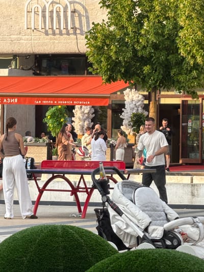 The image depicts an outdoor scene in front of a building with a red awning. A white and gray baby stroller is prominently positioned in the foreground. To the left, a woman with a braid, wearing white pants and a brown tank top, stands facing a red ping pong table. A man in a white t-shirt and dark pants is visible on the right, looking towards the left. In the background, several other individuals are present, some seated at tables and others standing. A large green tree with light green leaves shades part of the scene. The building facade above the awning features a decorative architectural element resembling three arches. The presence of Russian text on the awning suggests the location is in Russia, specifically Zelenograd, Russia. The overall atmosphere appears to be a casual outdoor gathering or dining setting.