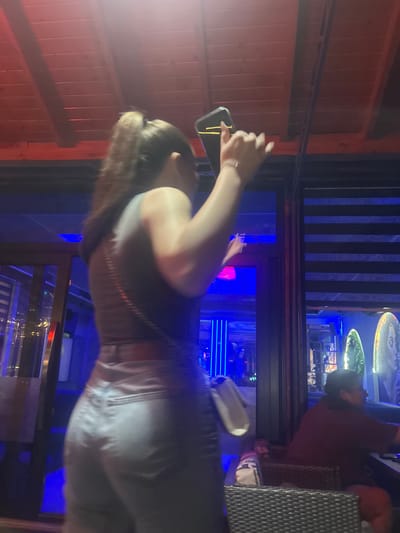 The image depicts an indoor night scene within an establishment located in Elhovo, Bulgaria. The ceiling is constructed from horizontal wooden slats, uniformly illuminated by a warm reddish-orange ambient light.
In the mid-ground, a female figure is positioned with her back to the viewer, slightly right of the frame's center. She has light brown hair styled in a high ponytail. She is wearing a light-colored, possibly grey, sleeveless tank top and light-toned jeans, with visible stitching and back pockets. Her right arm is raised, holding a black smartphone horizontally, which emits a bright yellow/orange light from its screen. Her left arm is also raised and slightly bent. A thin, dark strap, possibly from a shoulder bag, crosses her back diagonally. A white, irregularly shaped object, possibly a small bag or bottle, hangs near her left hip.
The background is dominated by intense blue and indigo ambient lighting. To the immediate left of the female figure, a vertical dark beam and a reflective surface (likely glass) show blurred reflections of interior lights. Further right, behind another dark vertical beam and a window section with horizontal dark blinds, a male figure is seated in a dark, woven wicker-style armchair. He is seen in profile, facing left, wearing a dark red short-sleeved top. A table, also dark, is in front of him. Beyond the seated man, decorative elements include two illuminated, elongated, green, organic-shaped objects, and an oval-shaped artwork featuring a face, illuminated by a soft white light.