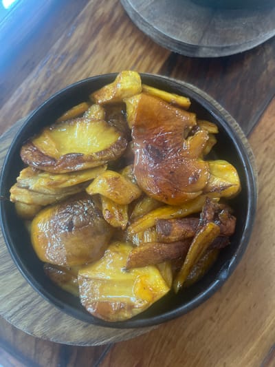 The scene in the city of Argveta, Georgia, depicts a dark, round skillet containing a cooked food item, likely root vegetables such as potatoes, presented on a wooden surface. The food consists of numerous irregularly shaped pieces, predominantly golden yellow to dark brown in color, exhibiting a glossy, caramelized surface texture. Some pieces appear as slices, while others are wedges or irregular chunks. The skillet is partially visible, primarily its upper rim and interior, and rests upon a lighter-toned wooden surface with visible grain. A segment of a second, darker wooden object is partially visible in the upper right background. No people or active interactions are observed. The environment suggests an indoor setting, likely a dining establishment or kitchen.