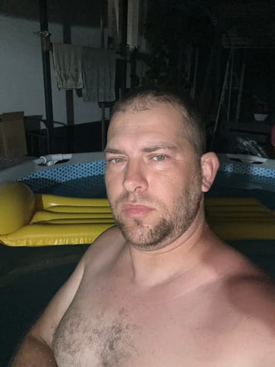 This media file shows a man, shirtless, from the chest up, in what appears to be a dimly lit outdoor setting at night. He has short, dark hair, a beard, and light-colored eyes. The man is positioned in the foreground, with his head tilted to his left. Behind him, to his right, is a large yellow object that resembles an inflatable pool float. To his left and further in the background, there are dark, indistinct structures and what seems to be part of a fence or railing. The scene has a dark and somewhat mysterious ambiance, suggesting it might be late evening or night. No specific activity or event is clearly depicted, beyond the man posing for the camera. There is no visible text in the image. The provided location context is Pîrlița, Moldova.