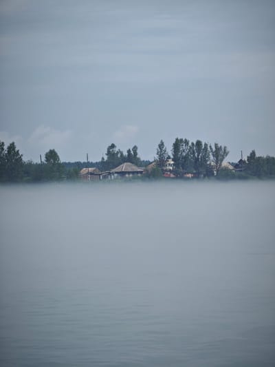 The image shows a body of water covered in a thick layer of fog. In the background, a line of trees and several rustic wooden houses are visible through the fog. The sky is overcast with a pale blue hue. The scene appears to be a rural or semi-rural landscape, possibly a village, as suggested by the presence of houses and trees. The location is identified as Atamanovo, Russia.