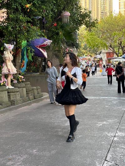 The image captures an outdoor setting likely in Baqiao, China. A young woman wearing a white cardigan, a black skirt, knee-high socks, and black shoes stands on a paved surface. In the background, multiple people walk in a crowded area that appears to be a market or public gathering. The environment includes trees and decorations; a mannequin in a pink dress lies on a stone surface near vegetation. A giant, artificial flower resembling a carnivorous plant is also visible. A tall building is present in the backdrop.