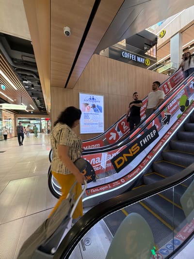 In Moskovskiy, Russia, a woman with dark hair, wearing a striped shirt and yellow pants, is seen on an escalator in a shopping mall. She is pulling a gray foldable shopping cart behind her. She is facing to her right and looking downwards. Other shoppers are visible on the escalator and in the background of the mall, which features a modern design with wooden accents and prominent advertising. One advertisement displays "DNS ГИПЕР" and another shows a clinic advertisement. A sign indicating "COFFEE WAY" is also visible above the escalator.
