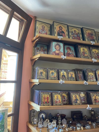 The image shows the interior of a shop selling religious artifacts in the city of Moni Archaggelou, Greece, at coordinates 40.597, 24.701. The shop is lined with wooden shelves that are full of framed religious icons. There are price tags on the shelves. Some of the icons are painted in a traditional Byzantine style, depicting saints and other religious figures. The shop also sells other religious items, such as candles, incense burners, and crosses. There is a large window in the background that looks out onto a courtyard.  The window frame is wooden, painted brown, and the walls are painted in a warm shade of light pink. The shelves are made of light wood, with a glossy finish. The icons are framed with simple wooden frames, also with a glossy finish.